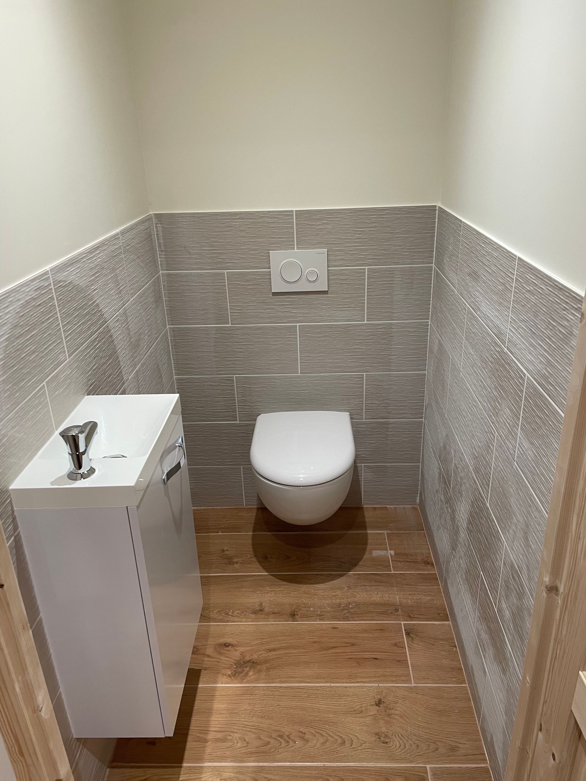 Une petite salle d'eau moderne avec des toilettes blanches suspendues, un carrelage au sol imitation bois et des carreaux muraux gris texturés.