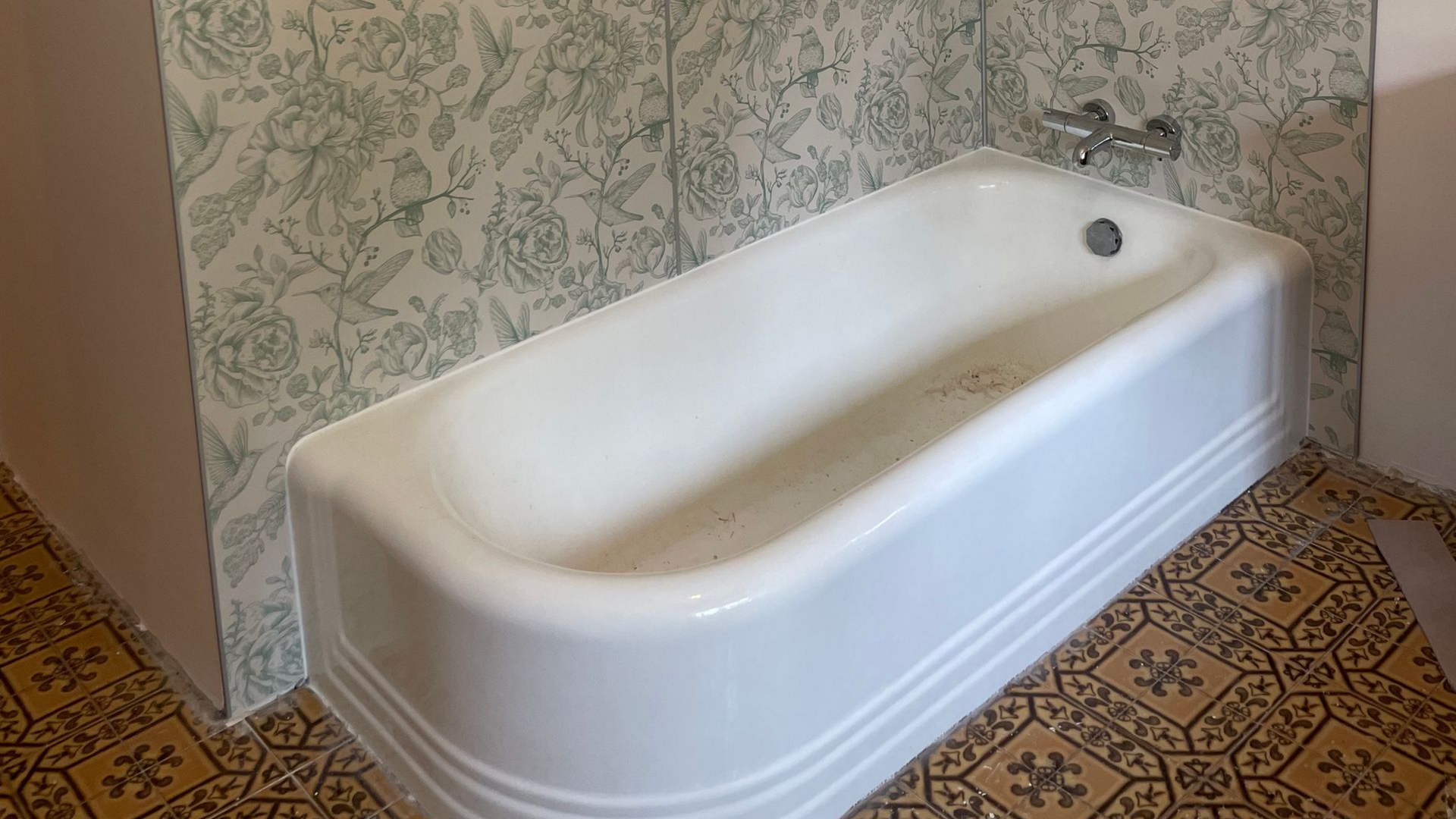 Une baignoire blanche est adossée à un mur tapissé de papier peint à motifs verts et blancs, sur un sol carrelé de tons beige et marron.