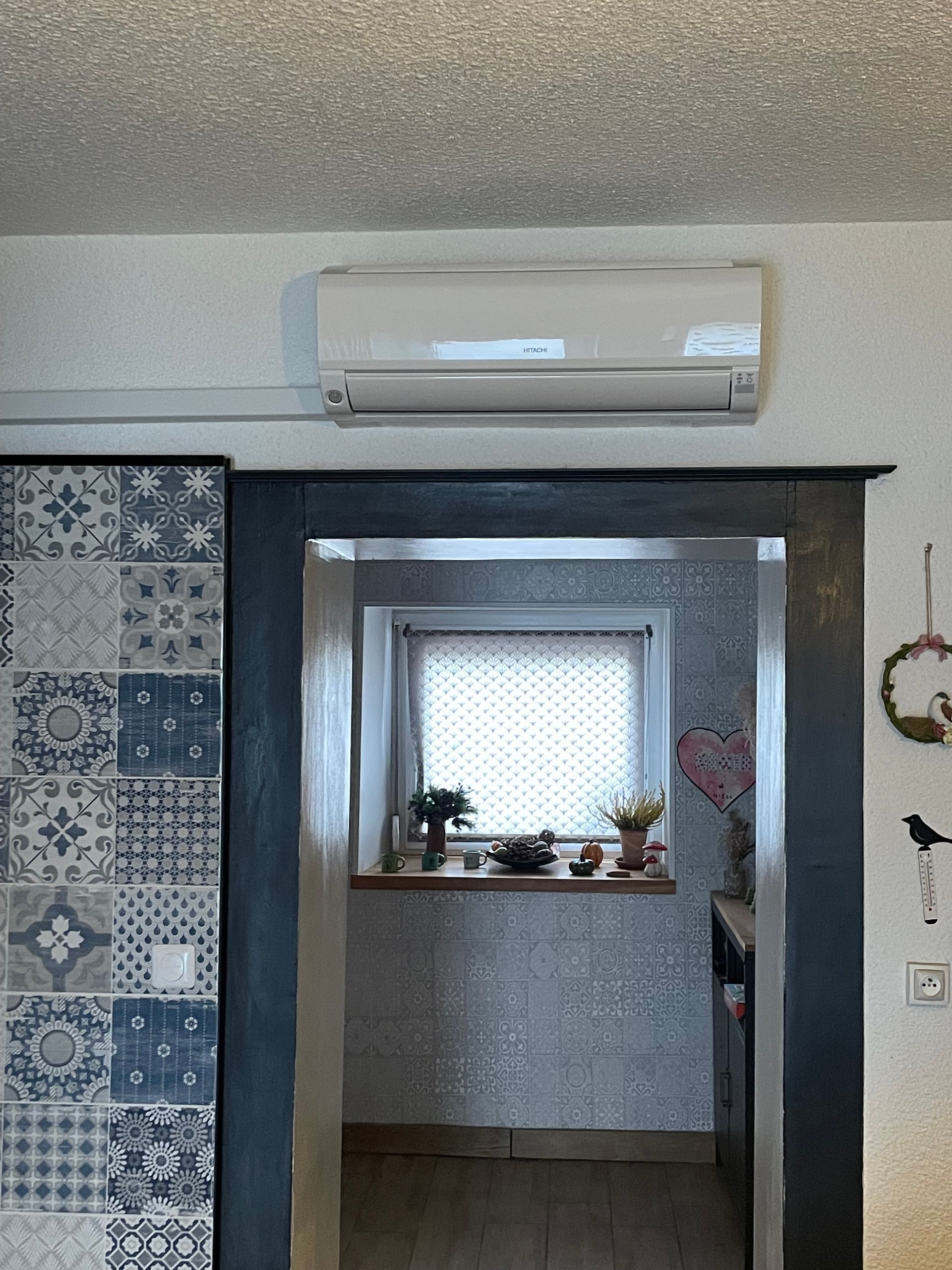 Un climatiseur mural blanc est installé au-dessus d'une porte donnant sur une pièce dotée d'une fenêtre et de panneaux décoratifs en carrelage bleu.