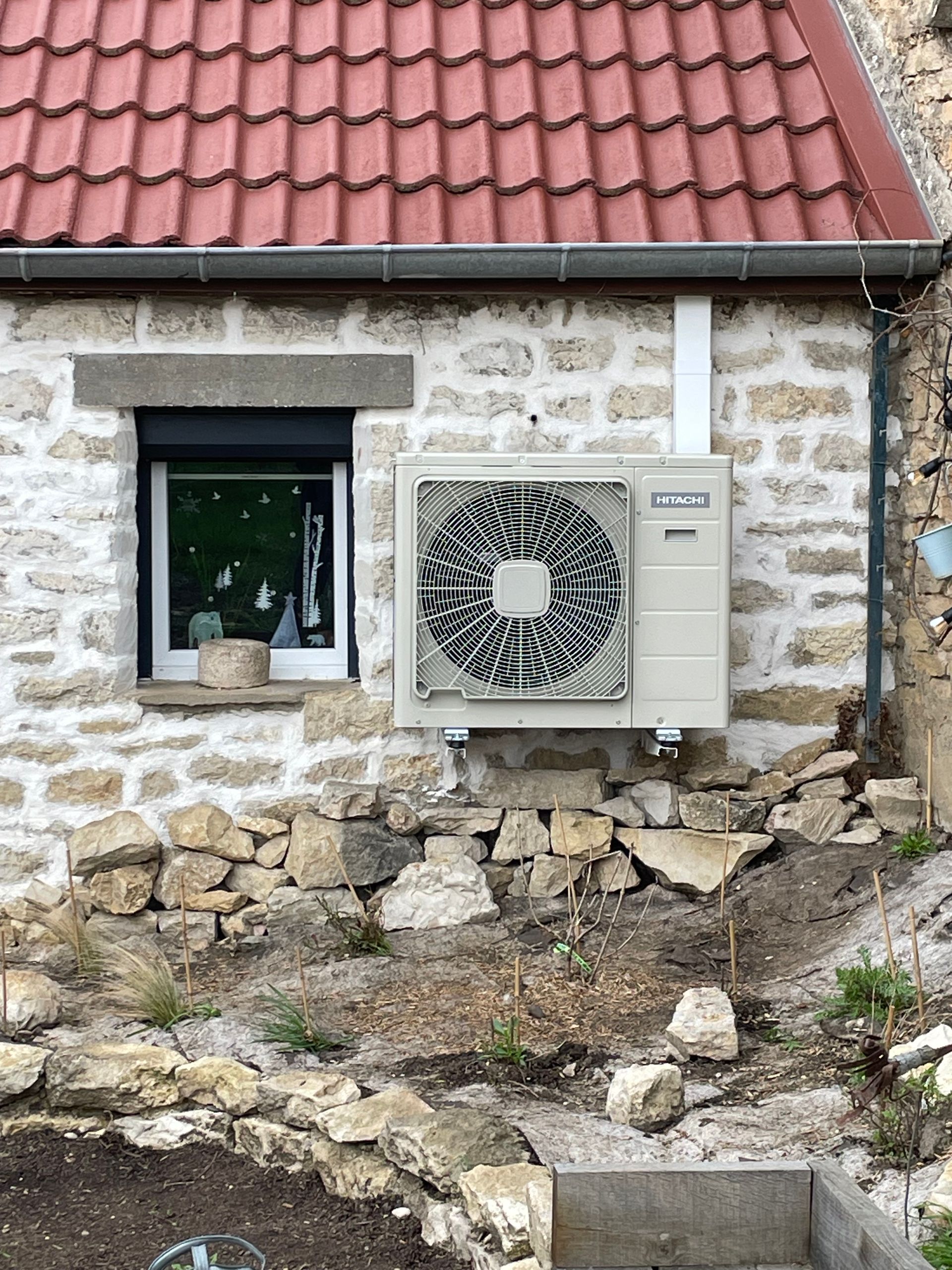 Une pompe à chaleur air-air beige est fixée sur le mur en pierre d'une maison au toit de tuiles rouges, à côté d'une petite fenêtre.