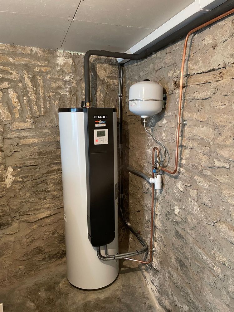 Un ballon d'eau chaude blanc avec un panneau de commande noir, à côté d'un vase d'expansion blanc, sur un mur de sous-sol en pierre brute.