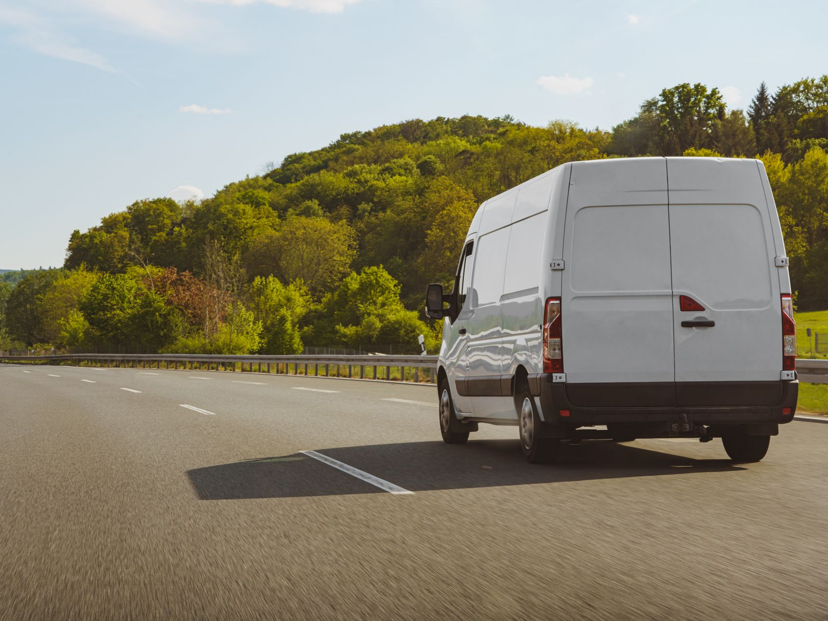 Une camionnette blanche circule sur une autoroute bordée de collines verdoyantes par une journée ensoleillée.
