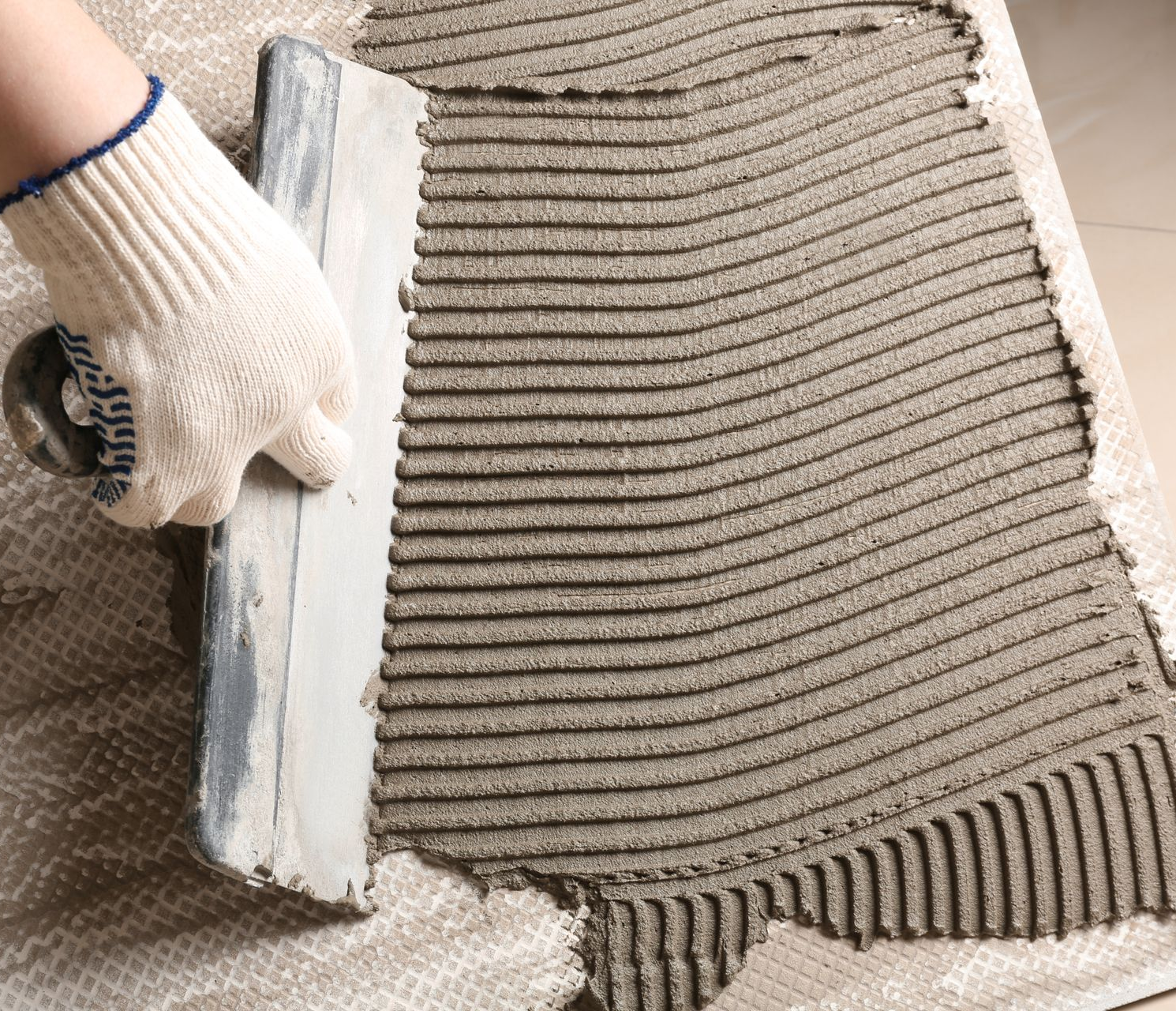 Main gantée utilisant une truelle crantée pour étaler du mortier gris sur une surface en vue de la pose de carrelage.