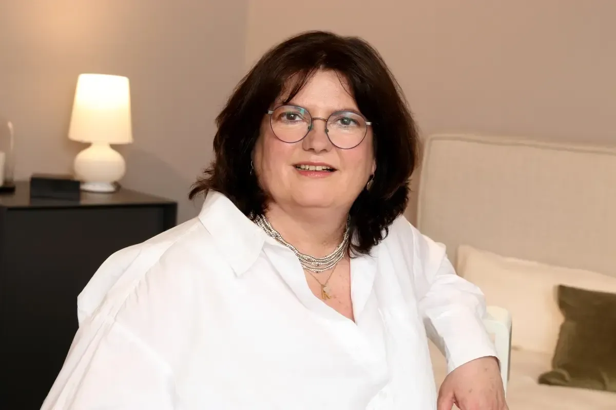 Eine Frau mit Brille und weißem Hemd sitzt auf einer Couch.