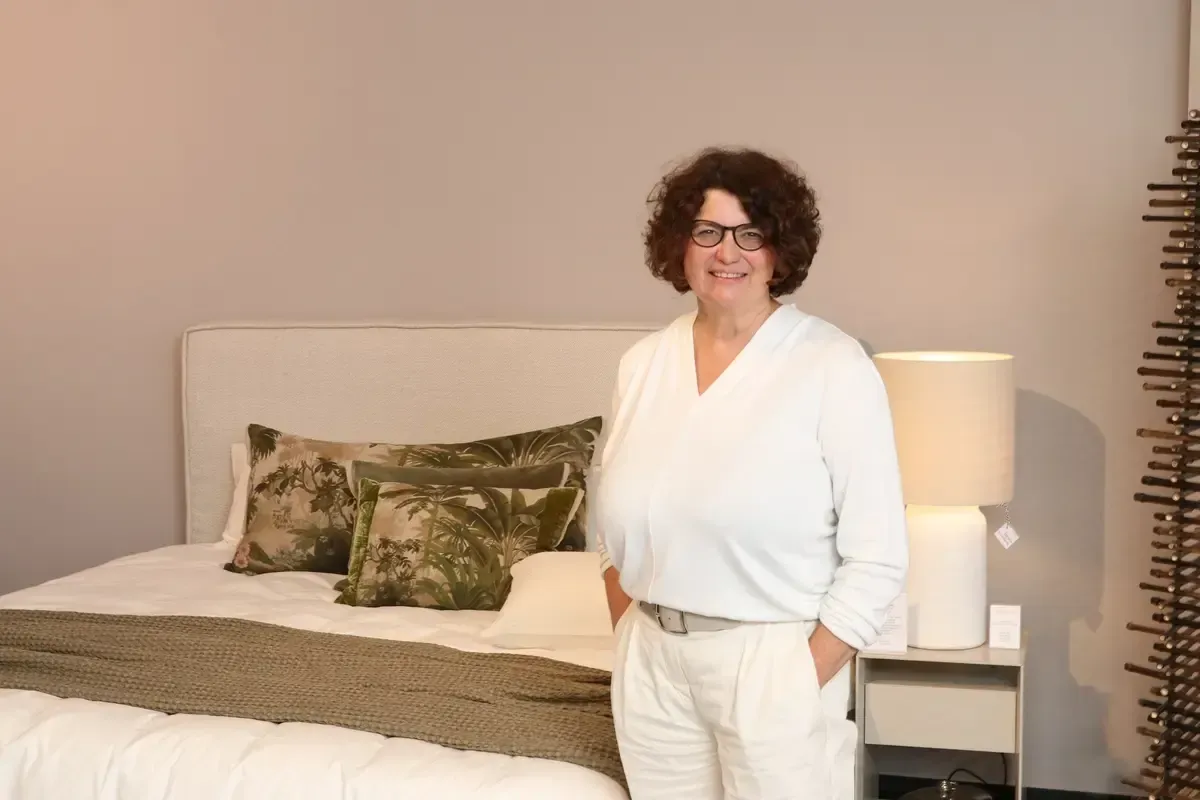 Eine Frau steht mit den Händen in den Taschen vor einem Bett.