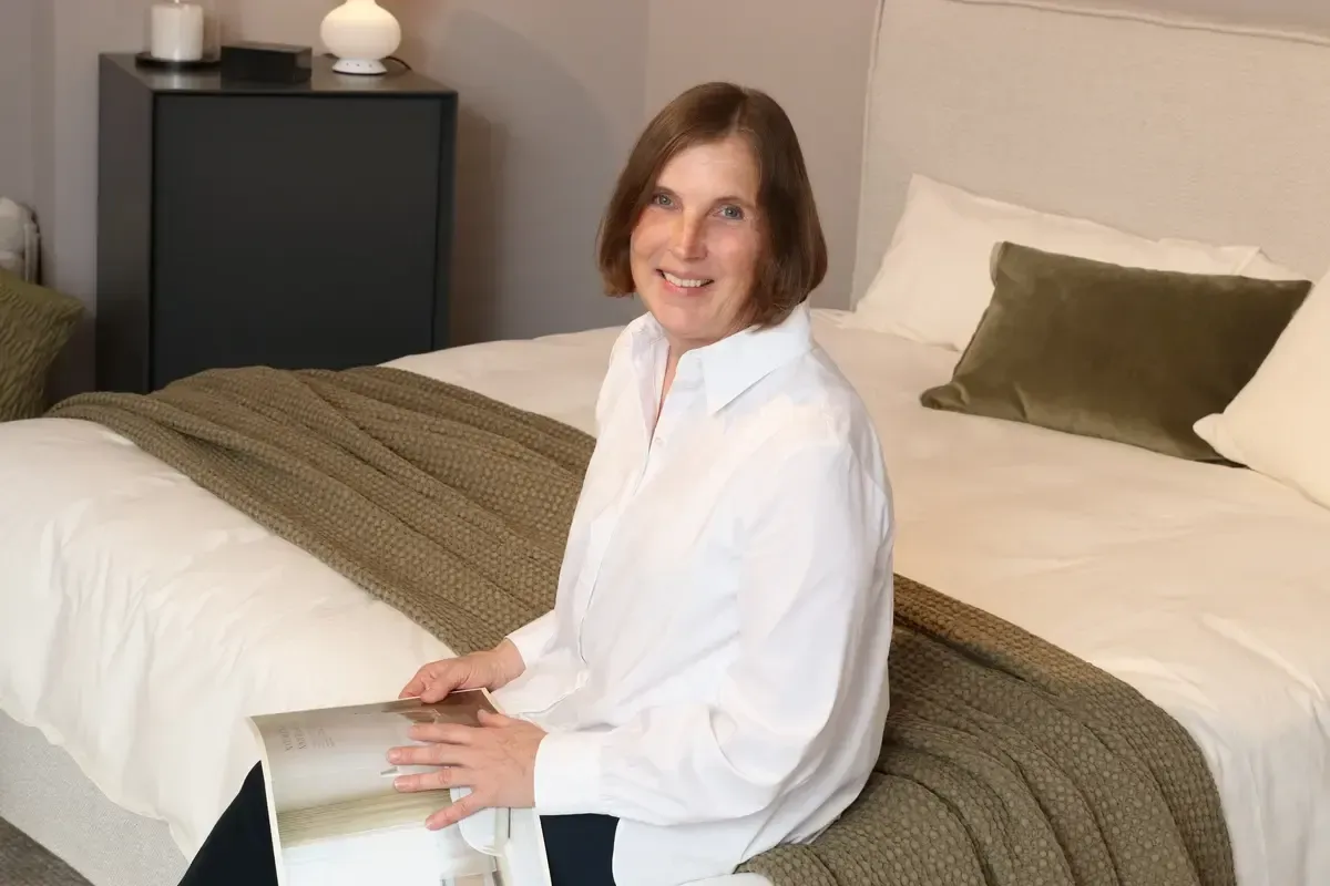 Eine Frau in einem weißen Hemd sitzt auf einem Bett und hält eine Zeitschrift in der Hand.