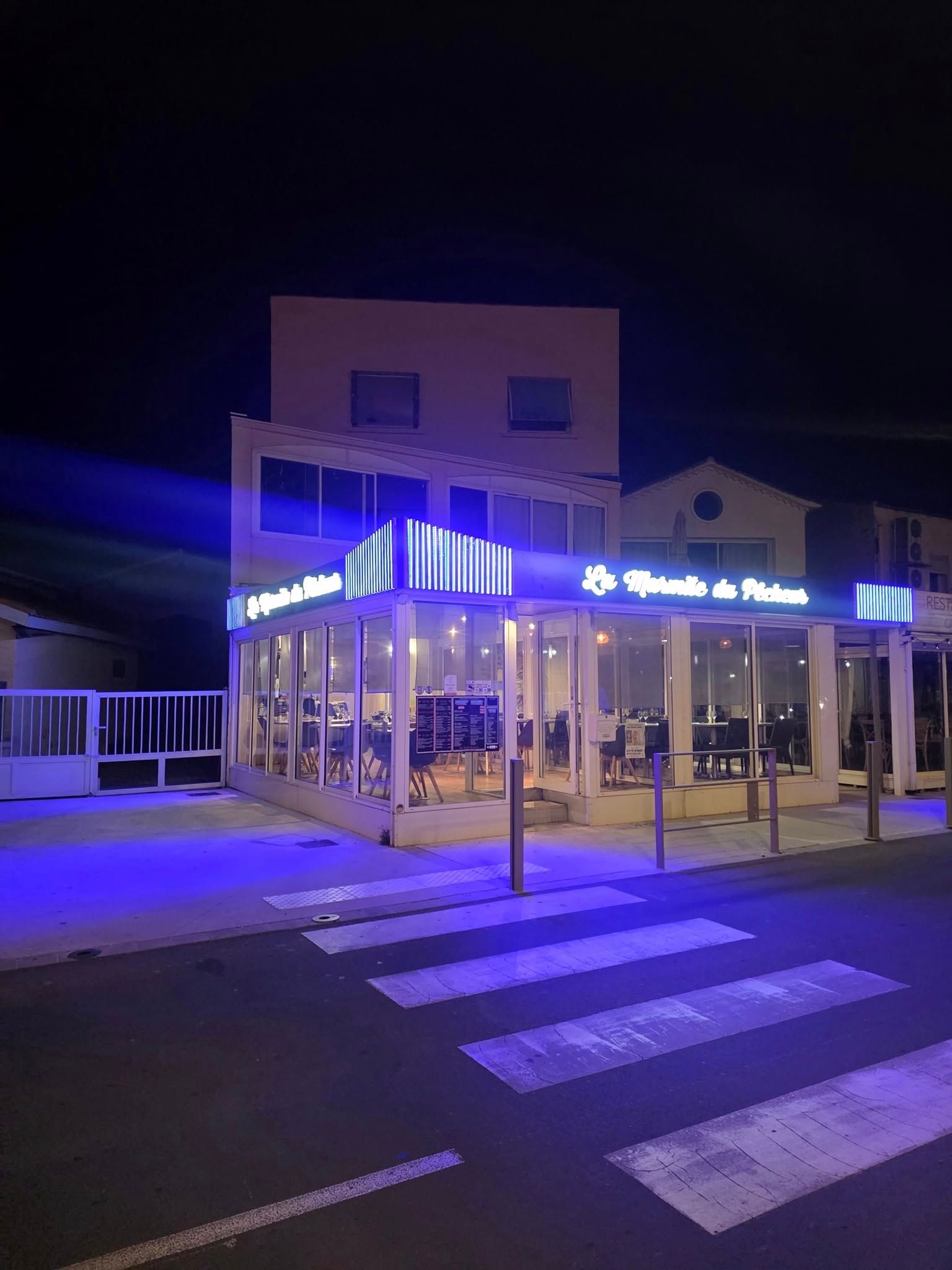 Un restaurant aux lumières vives, avec des cadres blancs et des enseignes au néon bleu la nuit, situé à côté d'une rue avec un passage piéton.