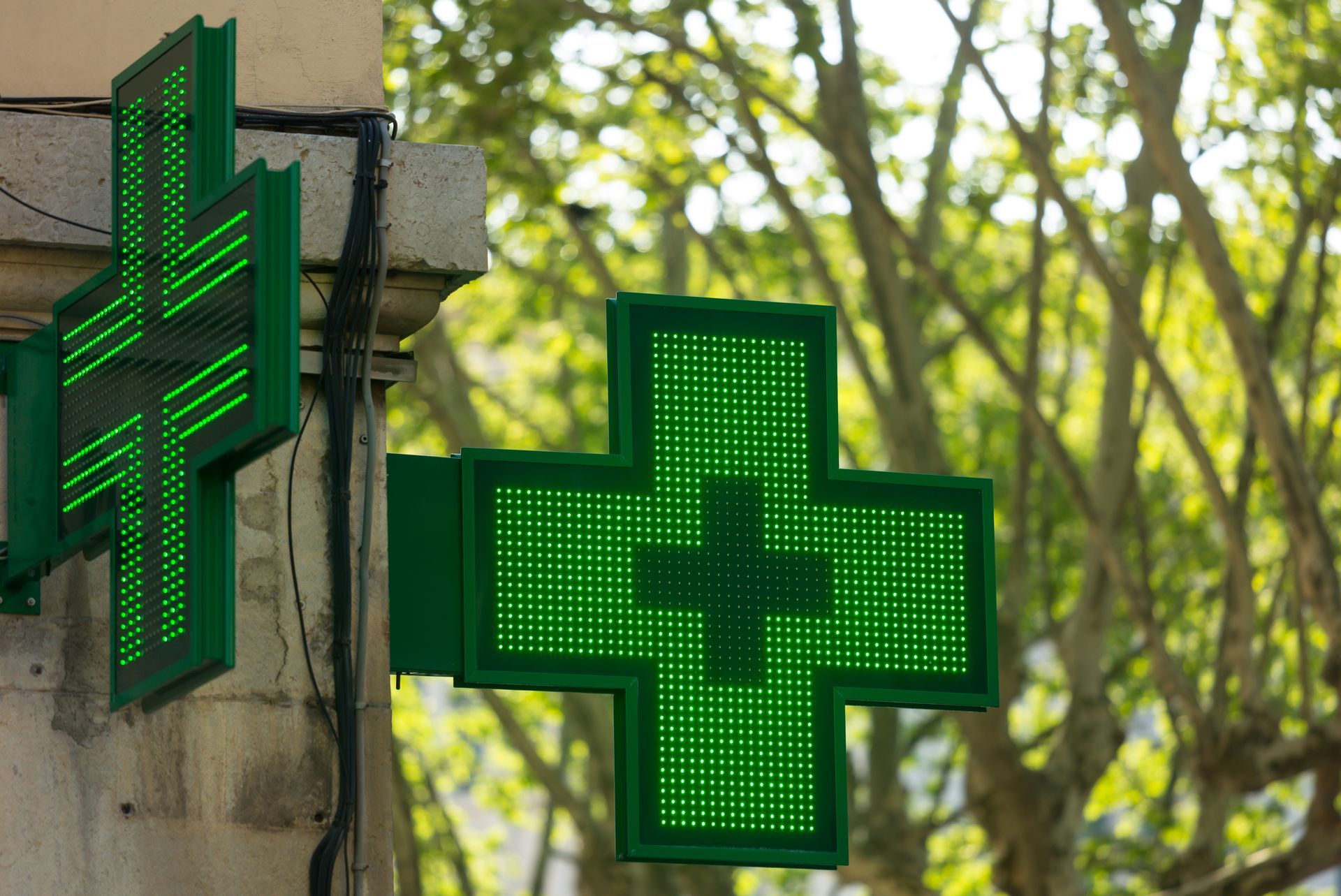 Deux enseignes en forme de croix, illuminées en vert, devant une pharmacie, sont fixées sur un bâtiment en pierre, avec des arbres flous en arrière-plan.