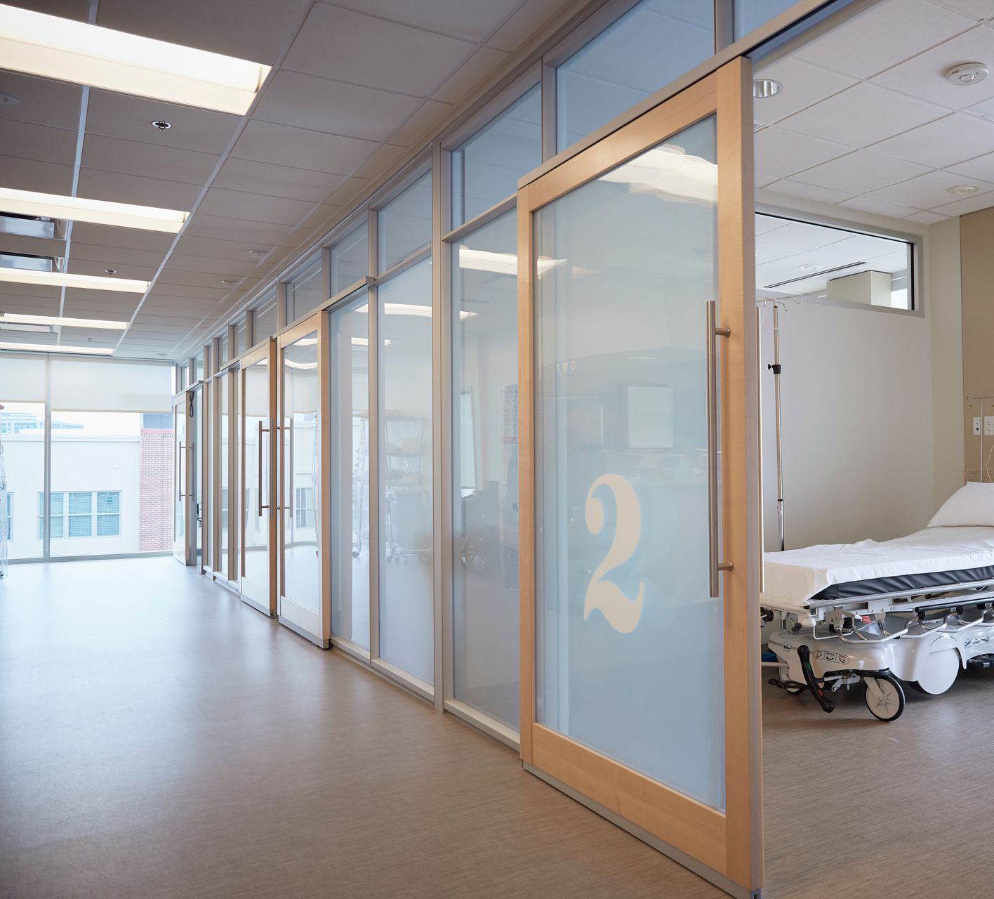 Un couloir d'hôpital avec des chambres de patients aux parois vitrées, dont une montrant un lit d'hôpital et le numéro 2 sur la porte.