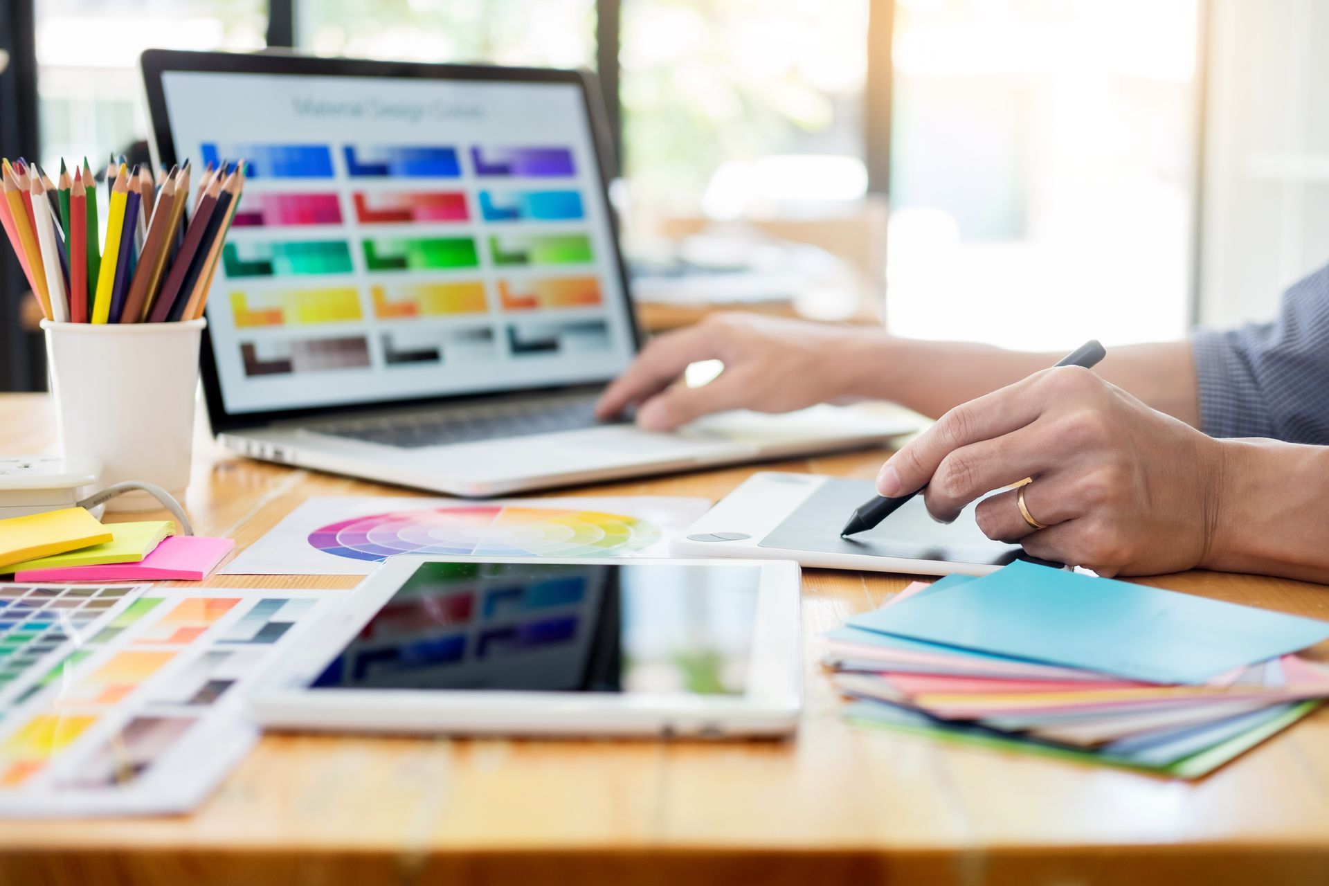 Une graphiste travaille sur une tablette numérique, un ordinateur portable affichant des palettes de couleurs, entourée de matériel artistique disposé sur un bureau.