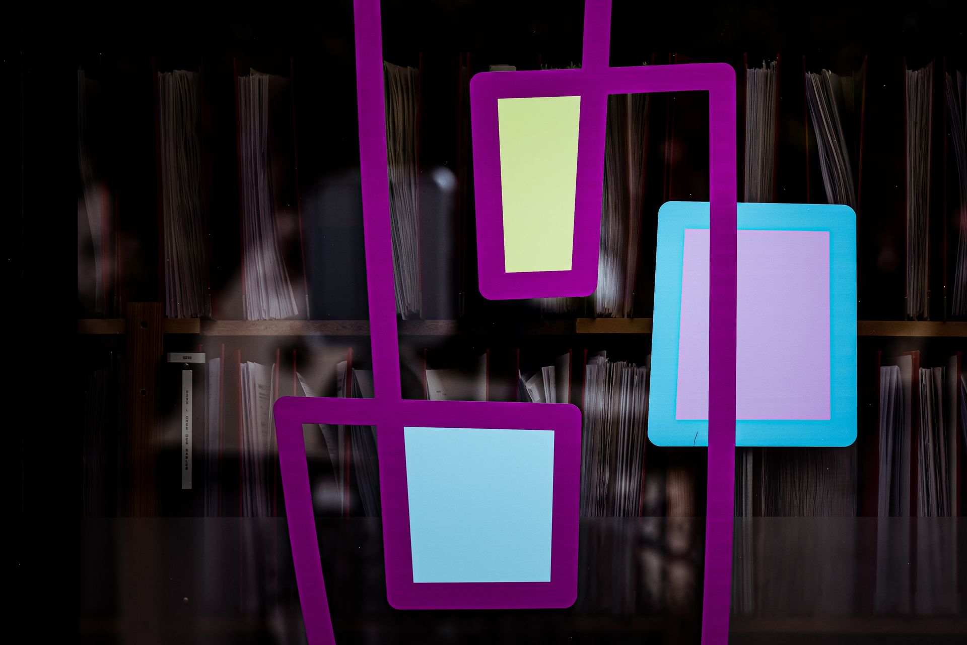 Un calque de dessin au trait violet représentant trois formes géométriques colorées sur un fond flou de livres empilés.