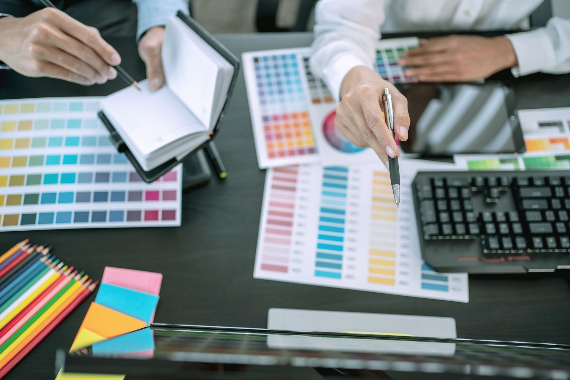 Deux personnes collaborent à un bureau avec des échantillons de couleurs, un carnet, un clavier d'ordinateur et des crayons de couleur.