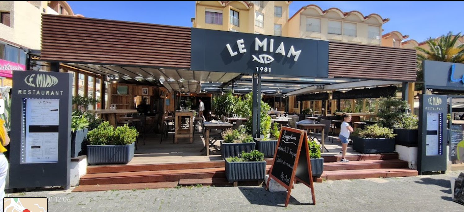 Entrée de la terrasse extérieure d'un restaurant avec l'enseigne LE MIAM, des éléments en bois, des plantes en pot et des tables par une journée ensoleillée.