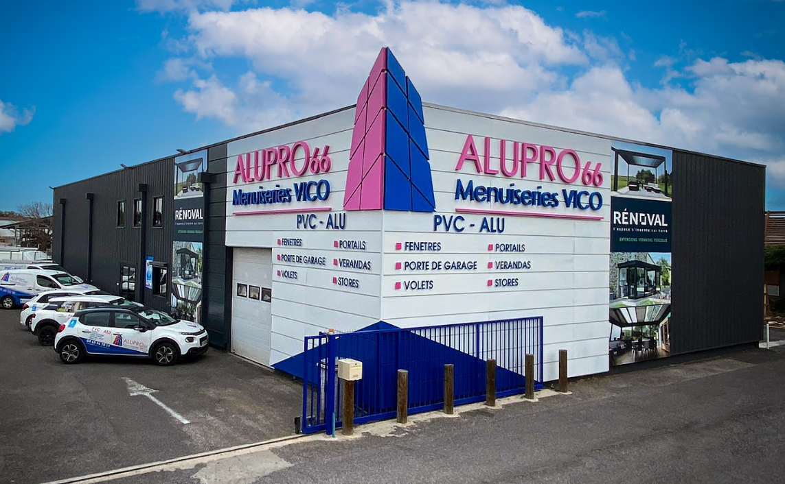 Vue d'angle d'un bâtiment commercial pour Alupro 66, avec des murs blancs, des accents bleus et une liste des services PVC/Alu.