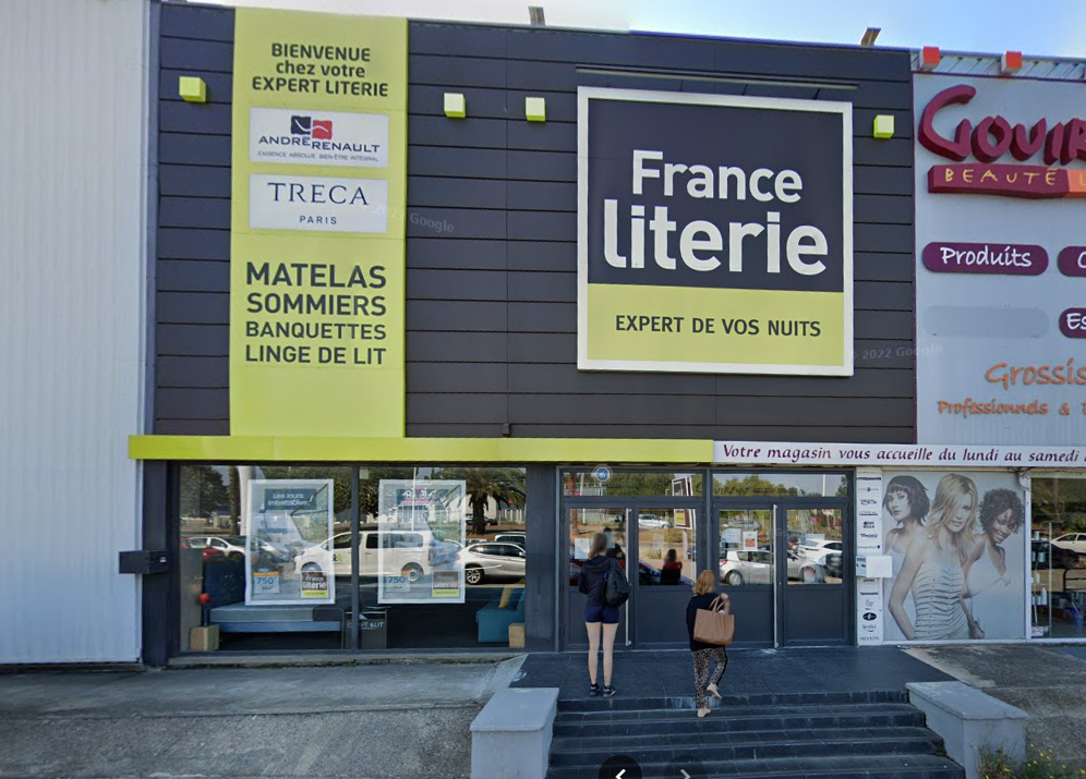 Deux personnes se dirigent vers l'entrée d'une librairie France Literie, reconnaissable à son enseigne jaune et noire, située dans une zone commerciale.