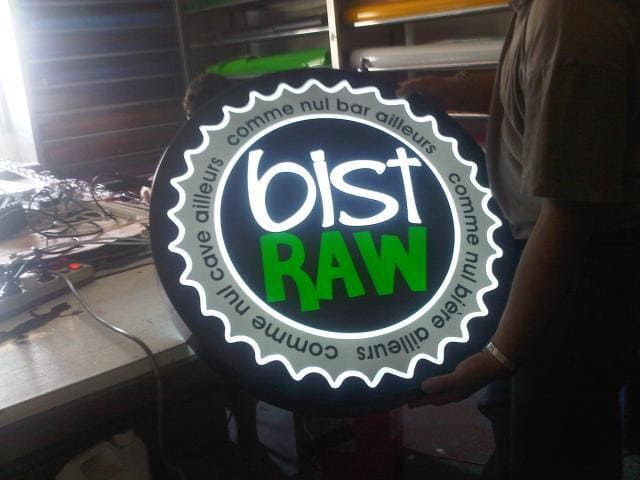 Une personne tient un panneau circulaire et lumineux sur lequel on peut lire bist RAW avec la phrase française comme nul bar ailleurs autour.