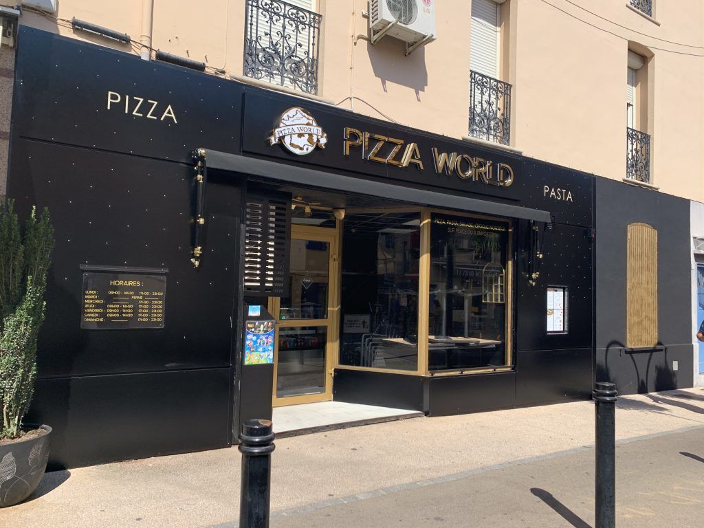 La façade d'un magasin Pizza World, avec des murs noirs, des lettres dorées et une entrée vitrée, située dans une rue ensoleillée.