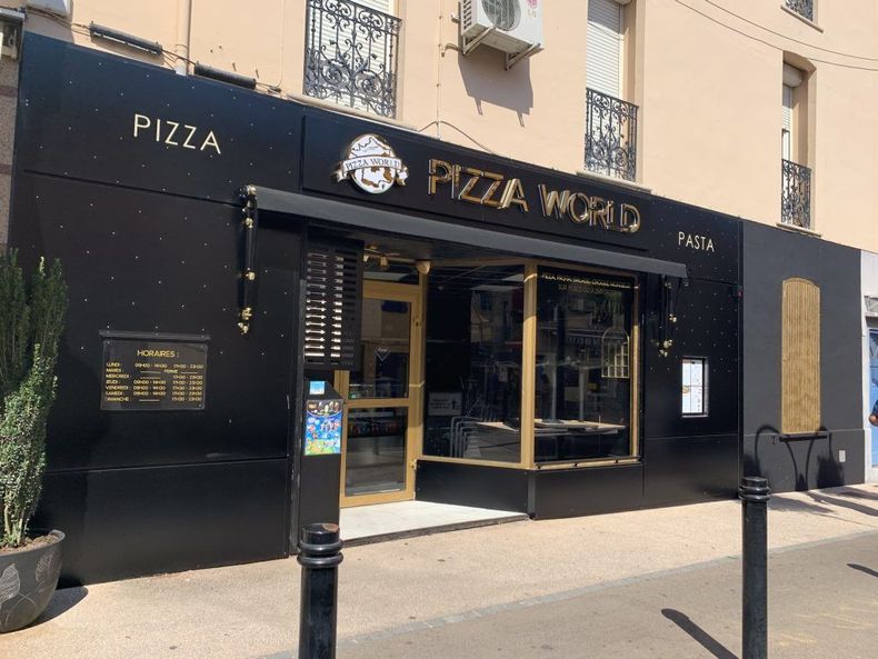 La façade d'un magasin Pizza World, avec des murs noirs, des lettres dorées et une entrée vitrée, située dans une rue ensoleillée.