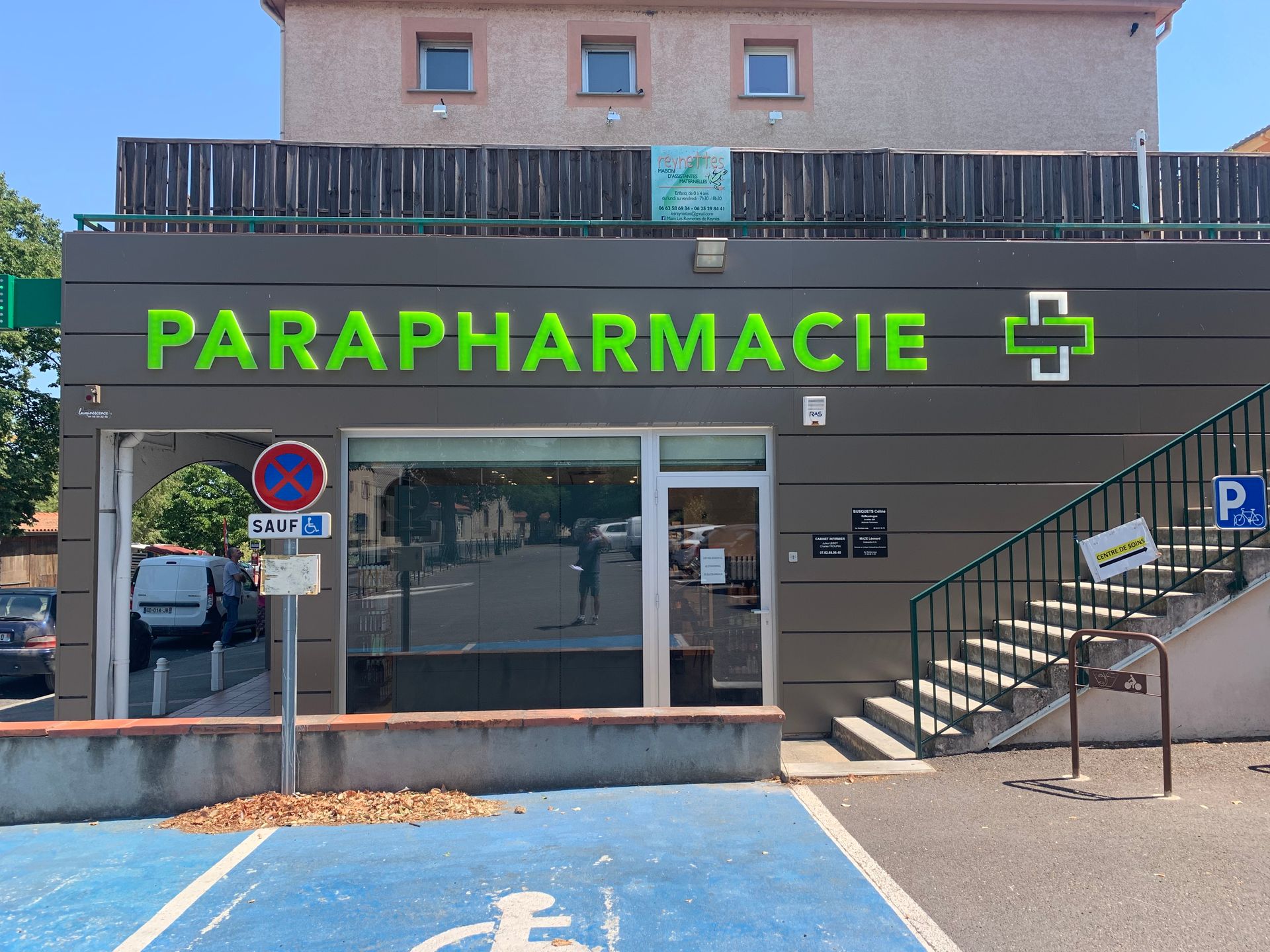Une façade de magasin avec une enseigne PARAPHARMACIE en lettres vert citron et un logo en forme de croix assorti, comportant des escaliers et une place de parking bleue.
