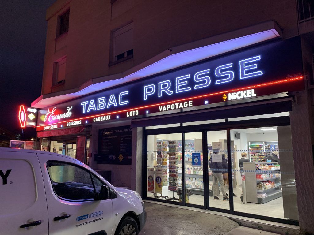 Vue nocturne d'une devanture de magasin avec une enseigne lumineuse TABAC PRESSE au-dessus de portes vitrées et une camionnette blanche garée.