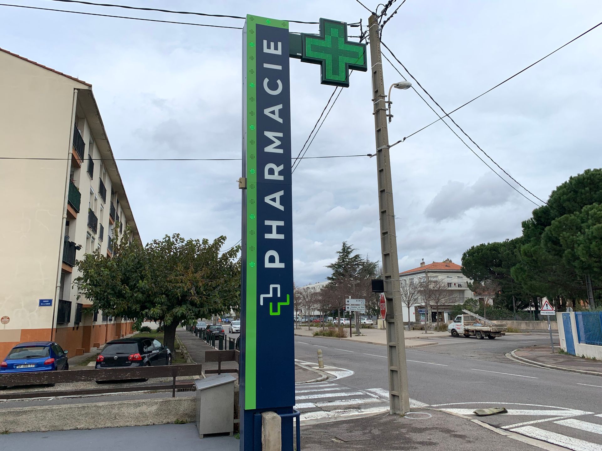 Une grande enseigne verticale bleue de pharmacie, ornée de touches de vert, se dresse sur un trottoir près d'une route et d'un immeuble de plusieurs étages.
