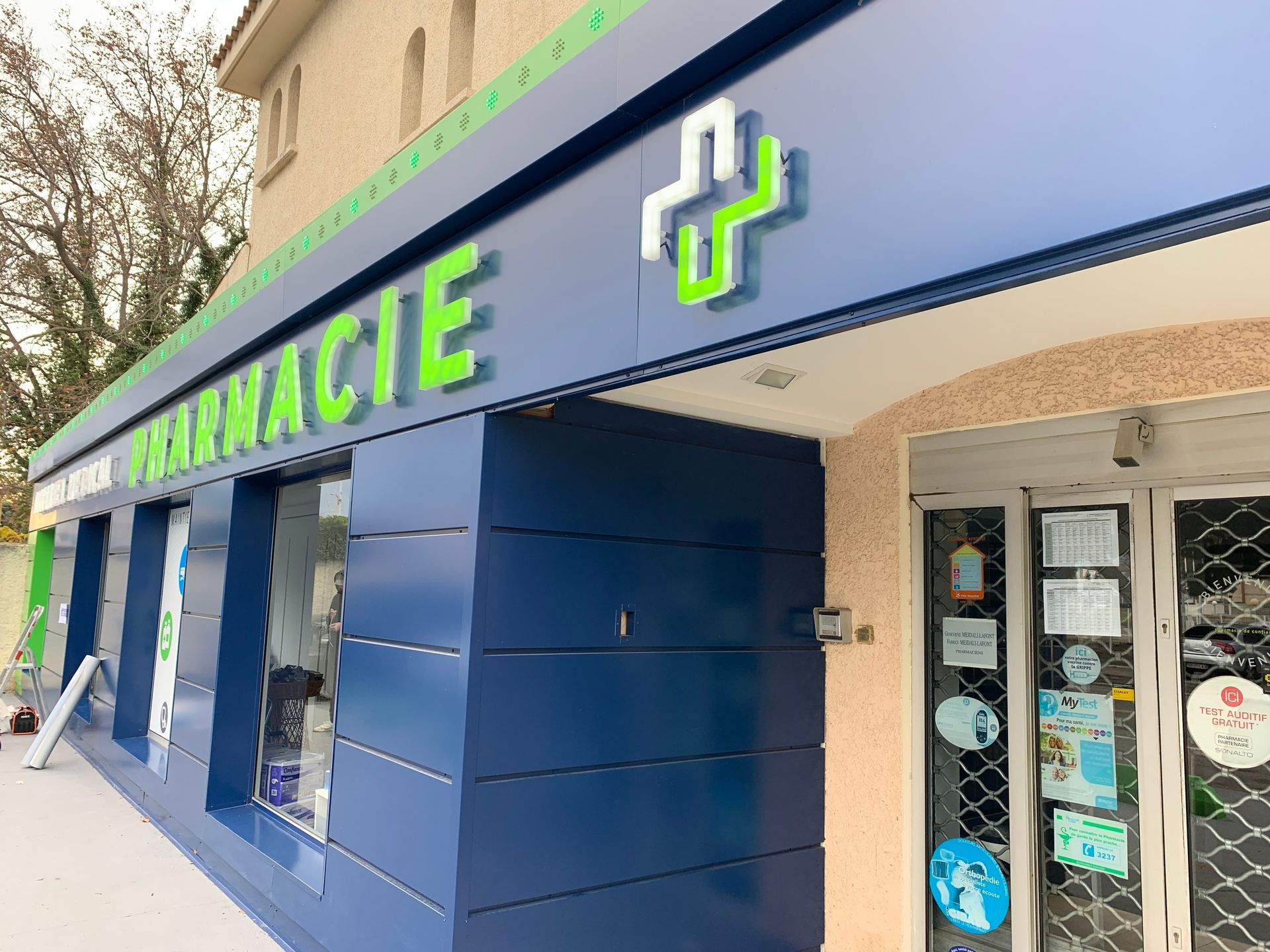 Une façade de pharmacie bleue avec une croix verte sur le mur et une grande enseigne verte PHARMACIE.