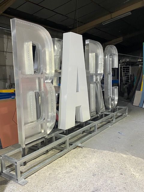 Grandes lettres 3D inachevées formant le mot BARC, montées sur une structure métallique à l'intérieur d'un atelier.