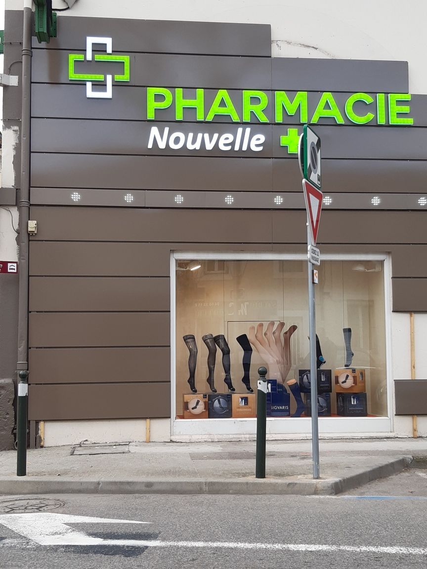 Façade marron de la Pharmacie Nouvelle avec une enseigne en forme de croix verte et une vitrine présentant des bas de contention.