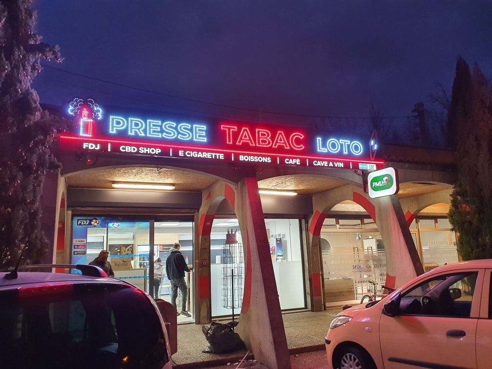La façade d'un magasin de tabac et de loterie, illuminée au crépuscule, présente des enseignes au néon et des voitures garées.
