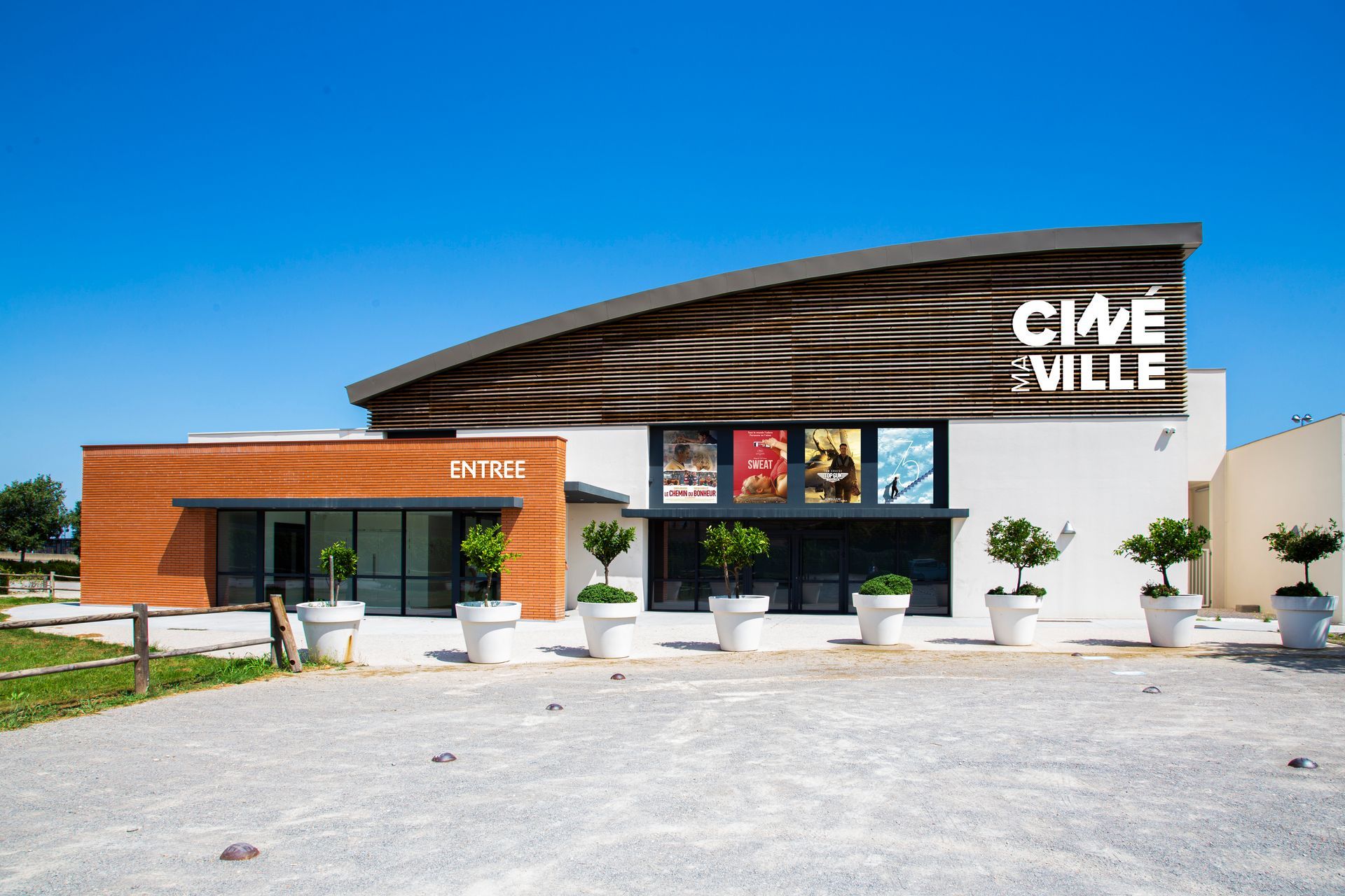 Un bâtiment de cinéma moderne nommé Cinéville, avec un toit en lambris, une entrée en briques et des arbres en pot devant.