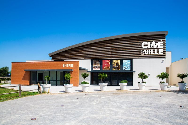 Un bâtiment de cinéma moderne nommé Cinéville, avec un toit en lambris, une entrée en briques et des arbres en pot devant.