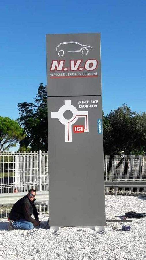 Un ouvrier est agenouillé près d'un grand panneau gris pour NVO et Decathlon, représentant un plan de rond-point sur une surface de gravier à l'extérieur.