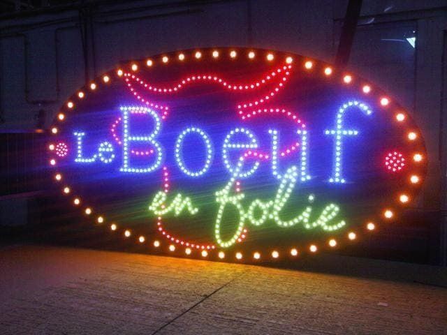 Un panneau ovale à LED affiche les mots Le Boeuf en folie en bleu et vert, entourés d'un contour de taureau rouge.