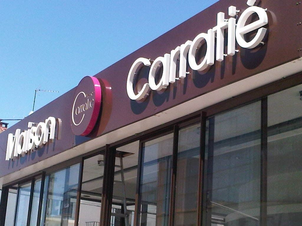 Une enseigne marron devant un magasin affiche Maison et Carré en lettres 3D blanches, avec un logo circulaire violet entre les deux.