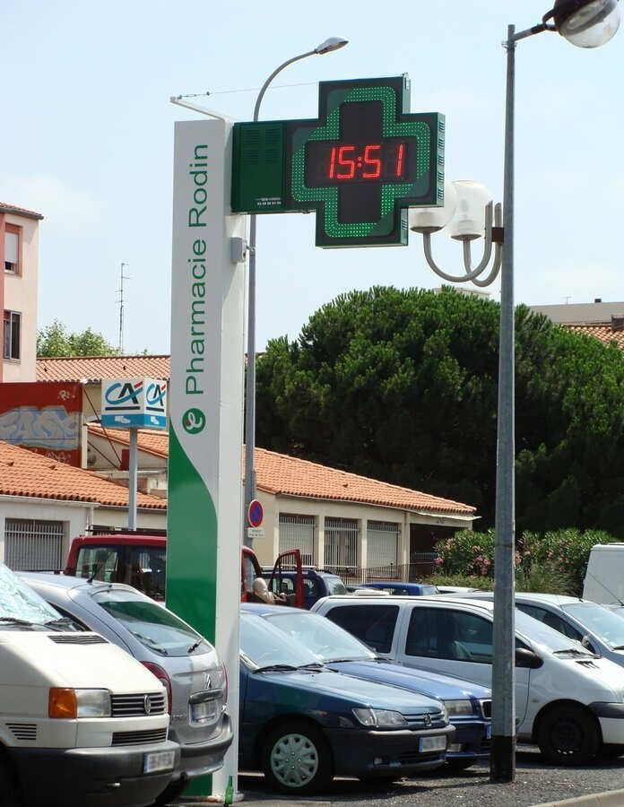 Enseigne de pharmacie avec une croix verte et une horloge numérique affichant 15h51, située sur un parking.