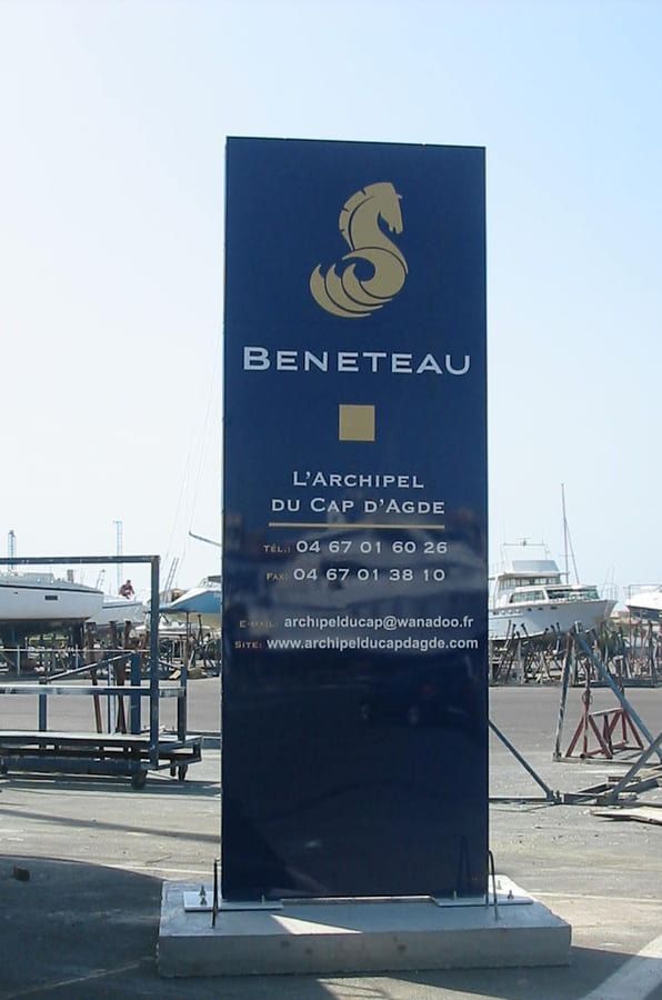Une enseigne rectangulaire bleu foncé pour Beneteau, L'Archipel du Cap d'Agde, se dresse dans un chantier naval avec des yachts en arrière-plan.