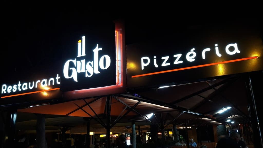 L'enseigne lumineuse du restaurant-pizzeria il Gusto la nuit, placée au-dessus d'une terrasse avec parasols.