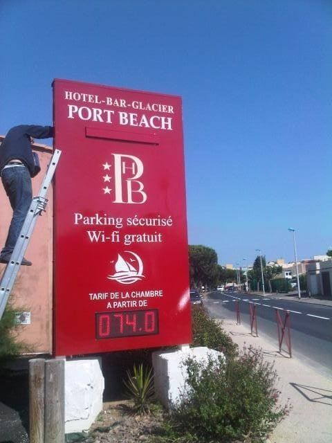 Une personne sur une échelle installe une grande enseigne rouge pour l'hôtel Port Beach, comportant un logo, la liste des équipements et un affichage des prix.