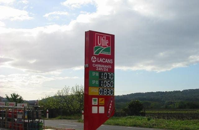 Un panneau rouge indiquant le prix du carburant dans une station-service, avec les logos Utile et Lacans sur fond de ciel nuageux.