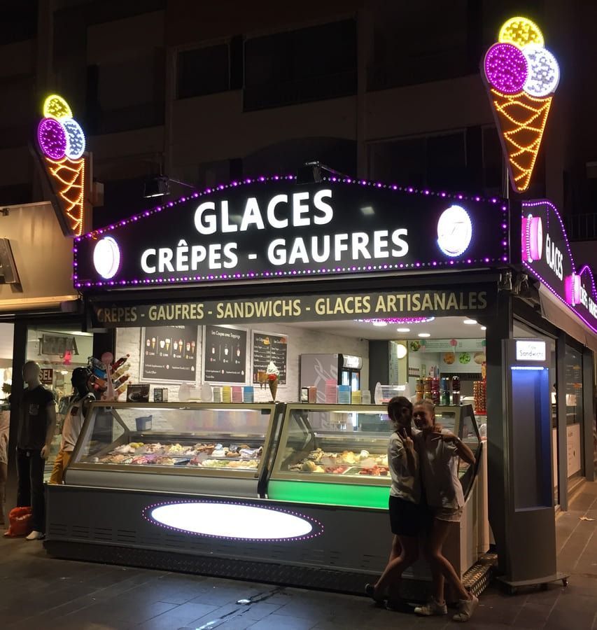 Deux personnes sourient devant un kiosque à glaces et à crêpes illuminé, de nuit.