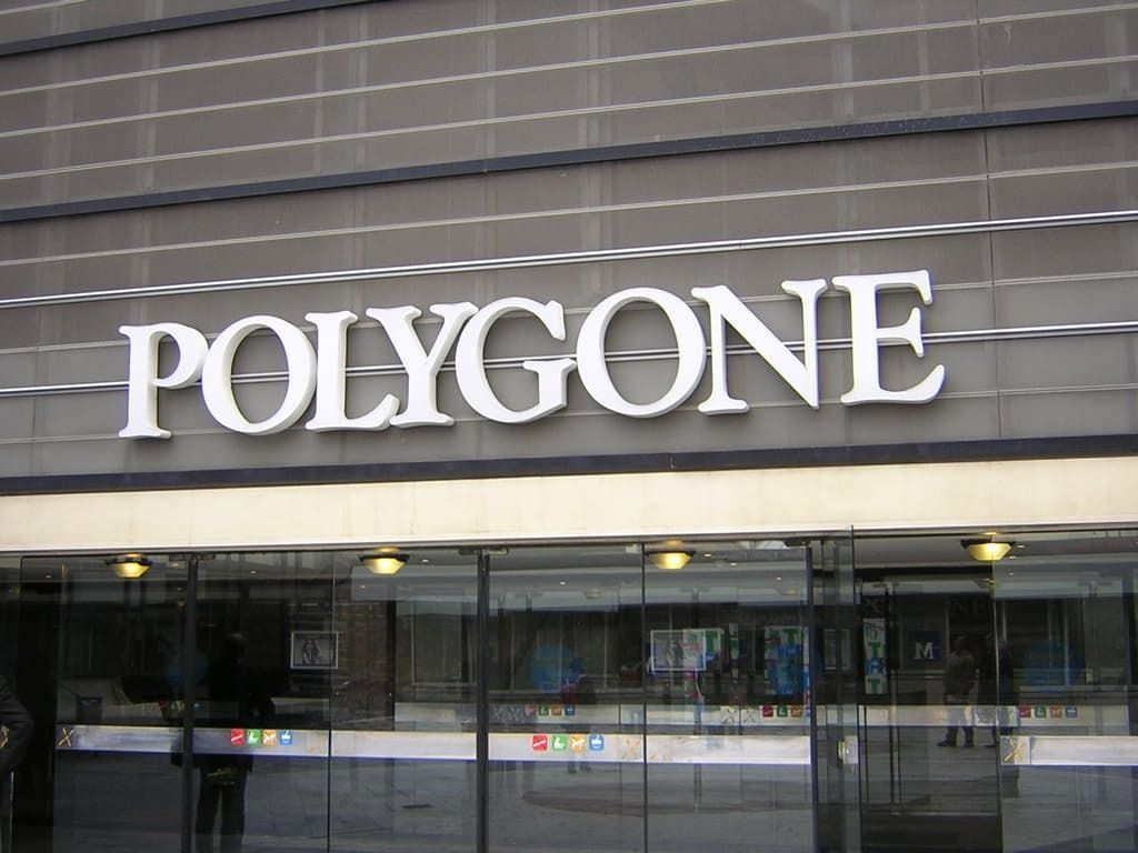 Le texte blanc à empattements POLYGONE est apposé sur la façade grise d'un bâtiment à panneaux horizontaux, au-dessus d'une vitrine.