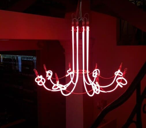 Un lustre moderne composé de tubes néon rouges incurvés et lumineux, suspendu dans une pièce sombre.