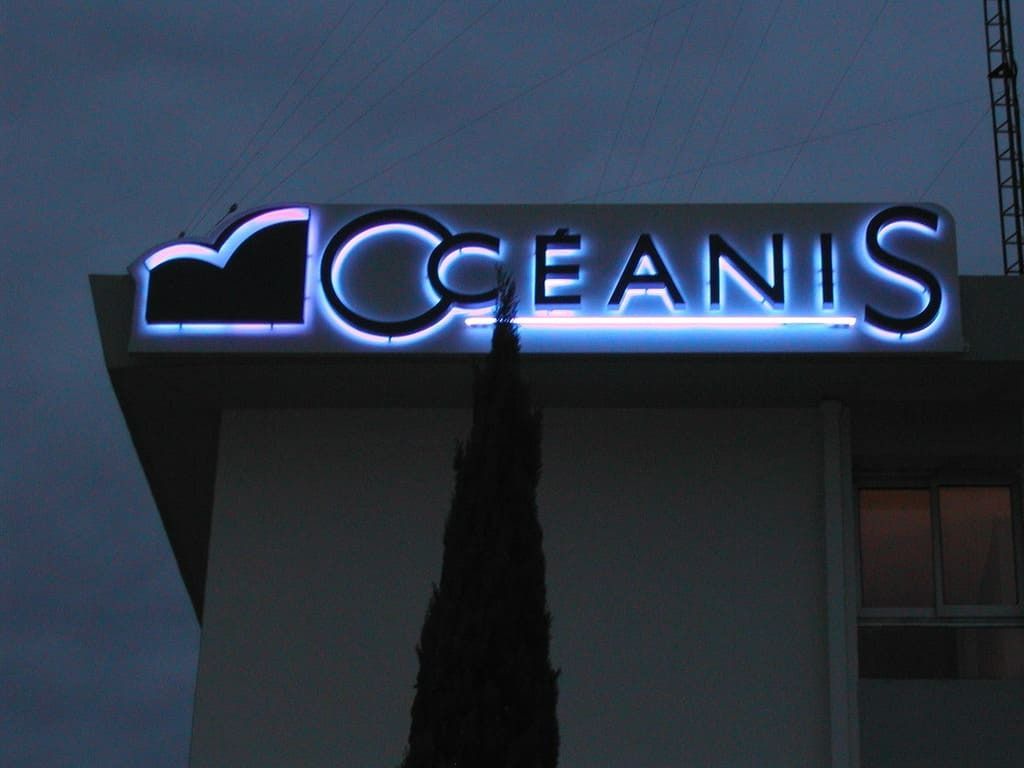 Une enseigne lumineuse blanche au néon, portant l'inscription OCEANIS, est fixée à l'angle d'un immeuble sur un ciel crépusculaire.