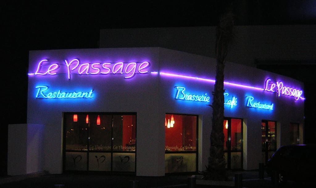 Un bâtiment de restaurant la nuit avec des enseignes lumineuses violettes et bleues indiquant Le Passage et Brasserie Café.
