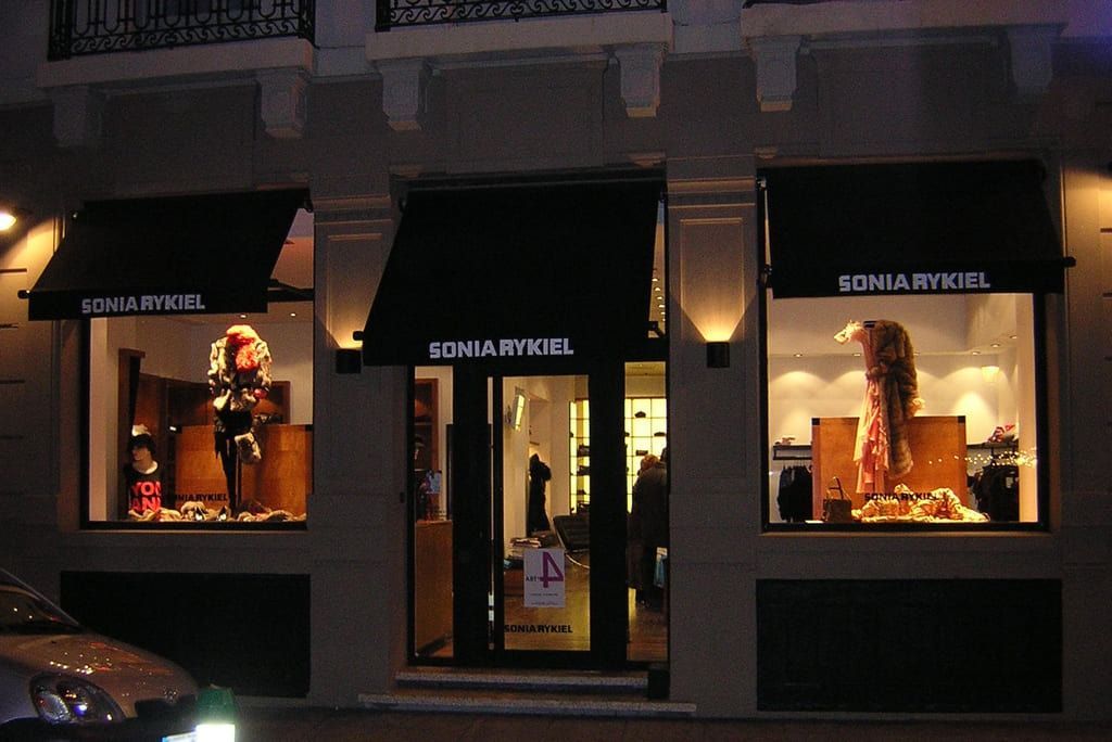 Une devanture de magasin avec trois auvents noirs au-dessus de grandes vitrines présentant des mannequins de vêtements, éclairées de l'intérieur la nuit.