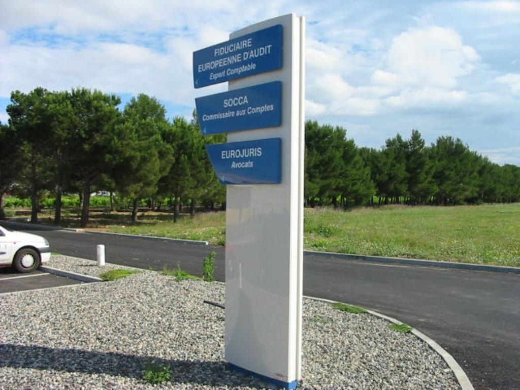 Un grand panneau vertical blanc, orné de trois pancartes bleues superposées, se dresse sur un terrain gravillonné, à côté d'une route goudronnée et de pins.