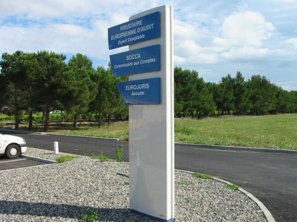 Un grand panneau blanc avec trois plaques bleues superposées répertoriant des entreprises, installé sur une zone de gravier au bord d'une route et d'arbres.