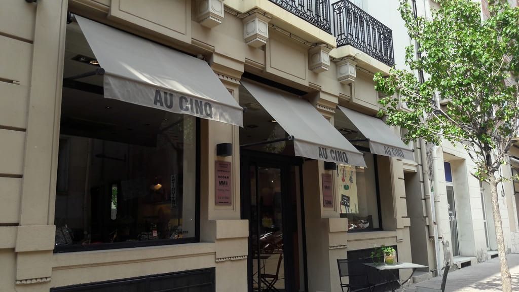 Vue depuis le trottoir de l'entrée d'un restaurant nommé Au Cino, avec des murs extérieurs beiges et des auvents gris.