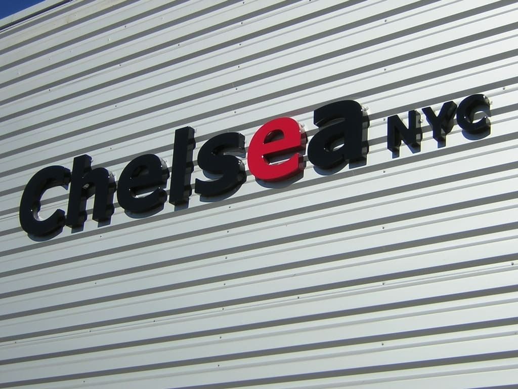 Le texte Chelsea NYC en lettres noires en relief sur un mur en tôle ondulée, avec la lettre e mise en évidence en rouge vif.