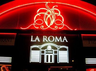 Une enseigne lumineuse au néon pour le restaurant La Roma, avec un texte blanc et une arche décorative rougeoyante la nuit.